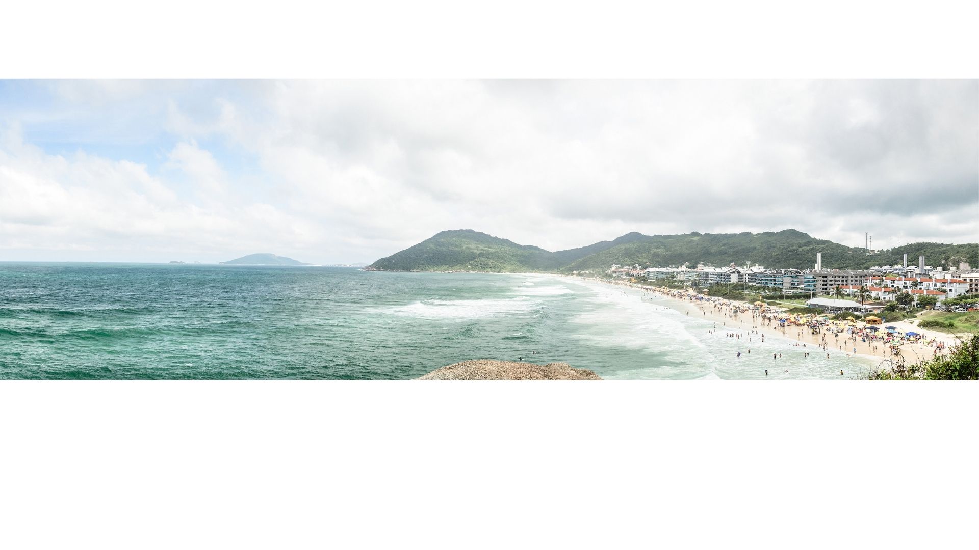 Vista panorámica de Praia Brava con olas, arena llena de bañistas y morros verdes al fondo