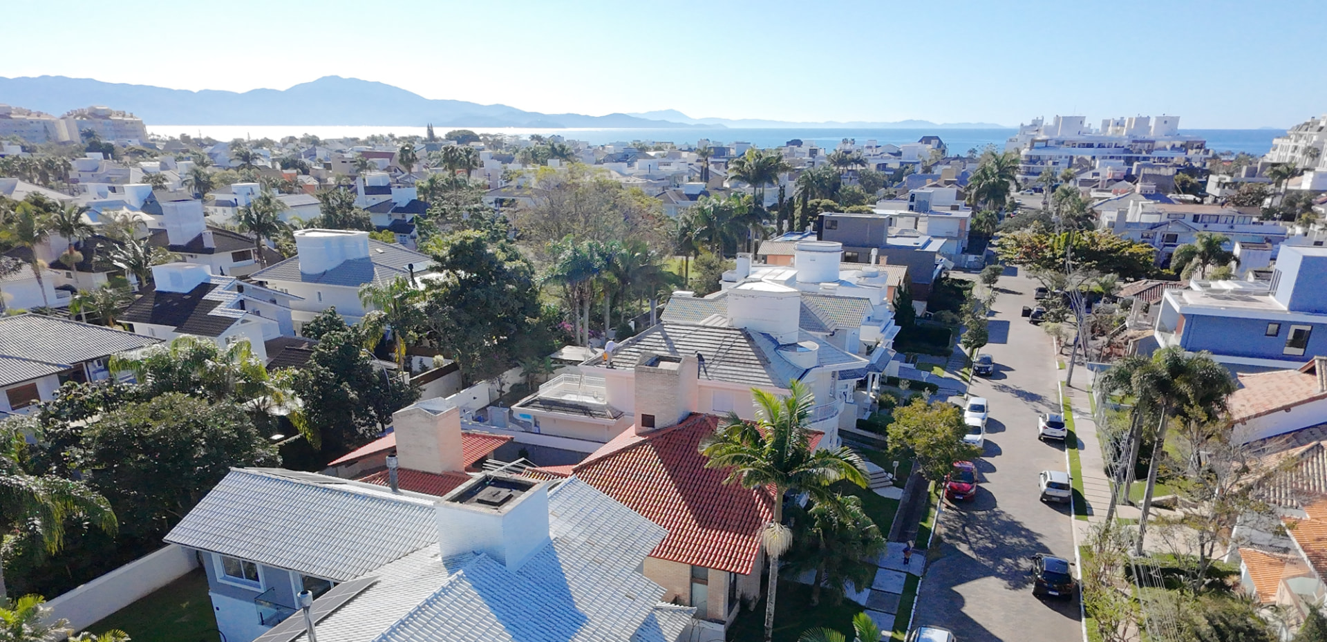 Vista aérea de un barrio residencial de Jurerê con casas bajas, palmeras, el mar y las montañas al fondo